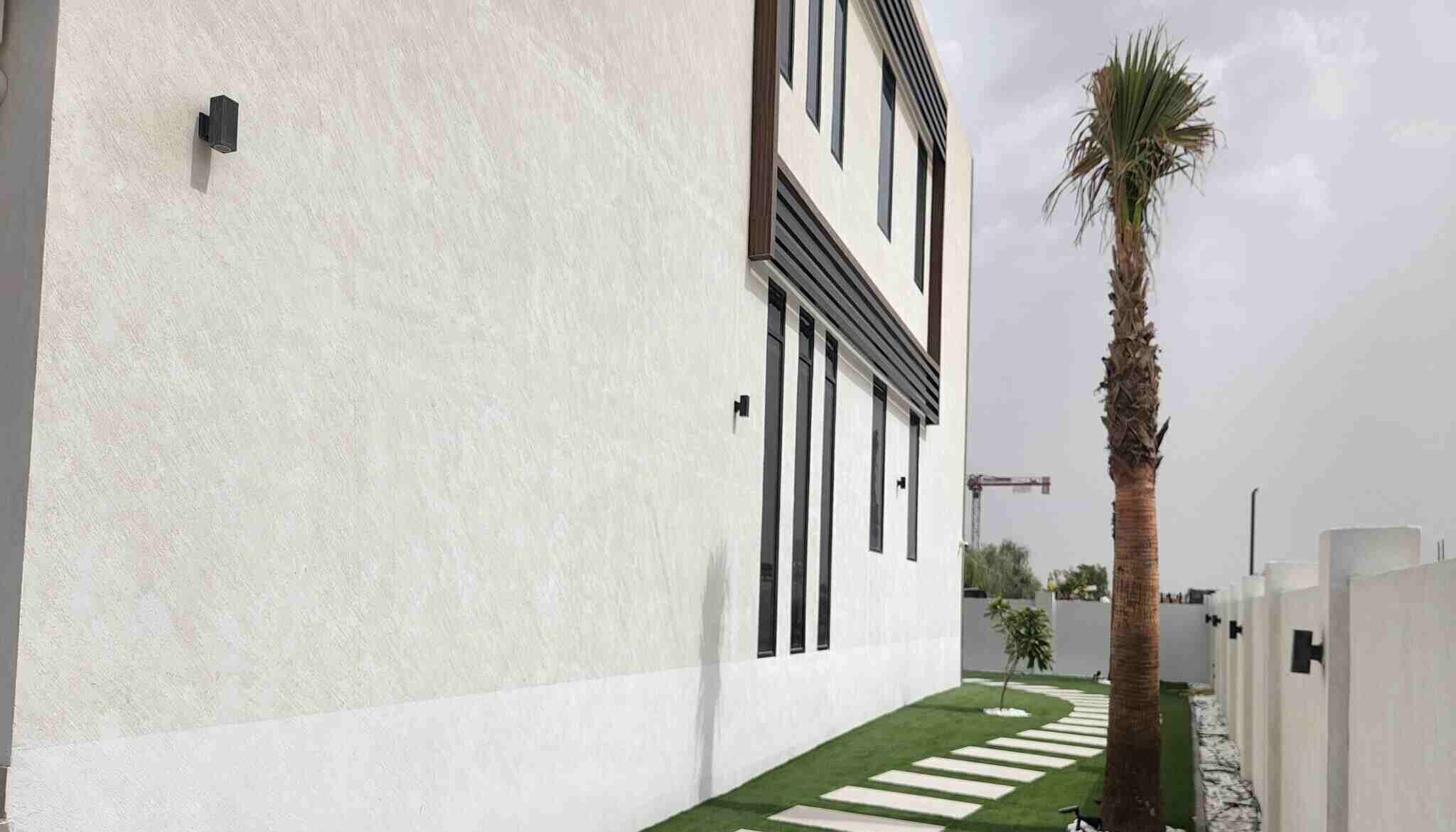 A beige building exterior with vertical windows on one side, a path of rectangular stones leading through a well-manicured lawn, and a tall palm tree on the right creates an oasis feel. Nestled in Dubai Hills, this residence epitomizes refined landscaping Dubai standards, all bordered by a white fence.