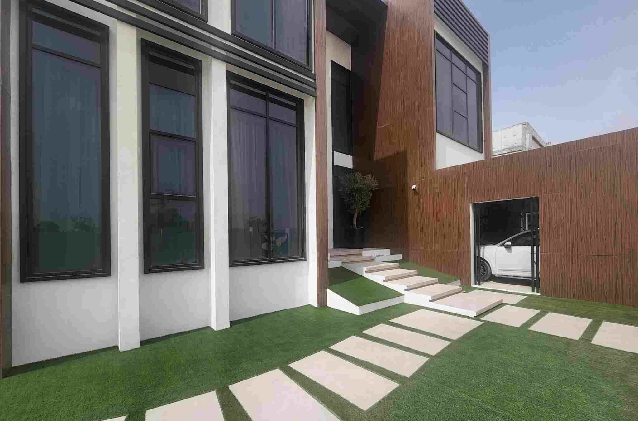 A modern house exterior in Dubai Hills boasts large black-framed windows, white and brown walls, and a landscaped lawn. Stone steps lead up to the entrance, with a tree nearby. A white car is parked in an open garage, showcasing the luxurious lifestyle that includes top-tier swimming pool services.