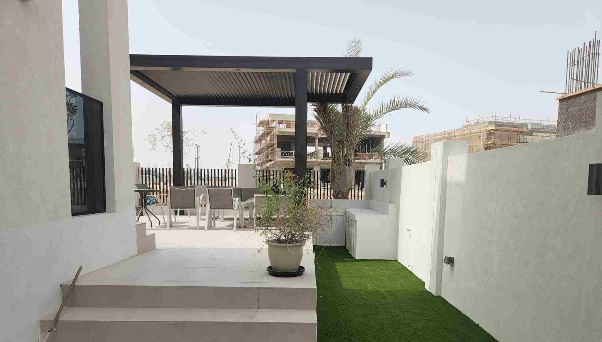 A modern outdoor area with a step leading up to a patio. The patio features a pergola with seating underneath, a potted plant on the step, and artificial grass lining the pathway. In the background, construction of nearby buildings in Dubai Hills is visible.