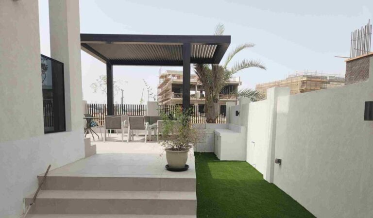 A modern outdoor area with a step leading up to a patio. The patio features a pergola with seating underneath, a potted plant on the step, and artificial grass lining the pathway. In the background, construction of nearby buildings in Dubai Hills is visible.