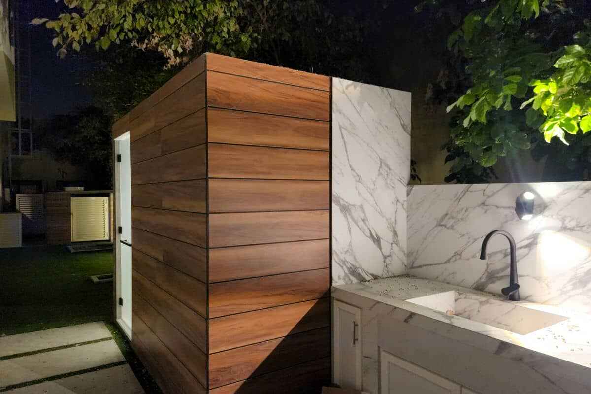 An outdoor kitchen at night features a white marble countertop with a sink and black faucet, illuminated by a wall light. Adjacent is a structure with horizontal wooden paneling. The backdrop includes trees and grass, perfect for those considering renovation or integrating Pool & Landscaping Dubai services