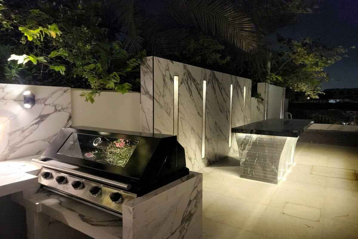 An outdoor kitchen at night with a modern marble countertop and backsplash. There's a covered grill on the left and another counter in the background. Soft lighting illuminates the area, highlighting the sleek and contemporary design, perfect for those who appreciate luxurious landscaping Dubai style