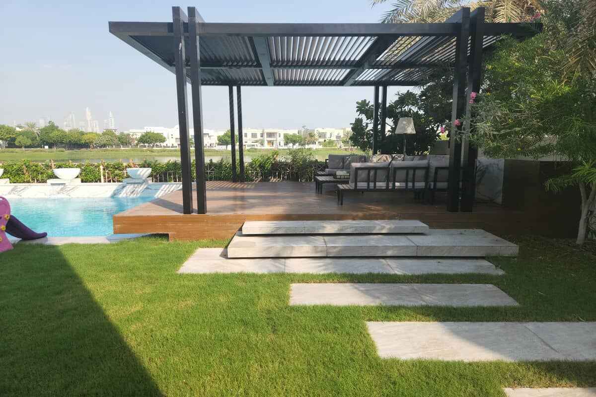 A backyard with a pool, wooden deck, and modern pergola. The pergola covers outdoor seating, providing shade. Stone steps lead from the green lawn to the deck. The area is beautifully landscaped with trees, shrubs, and distant views of neighboring houses—ideal for any pool renovation or landscaping Dubai project