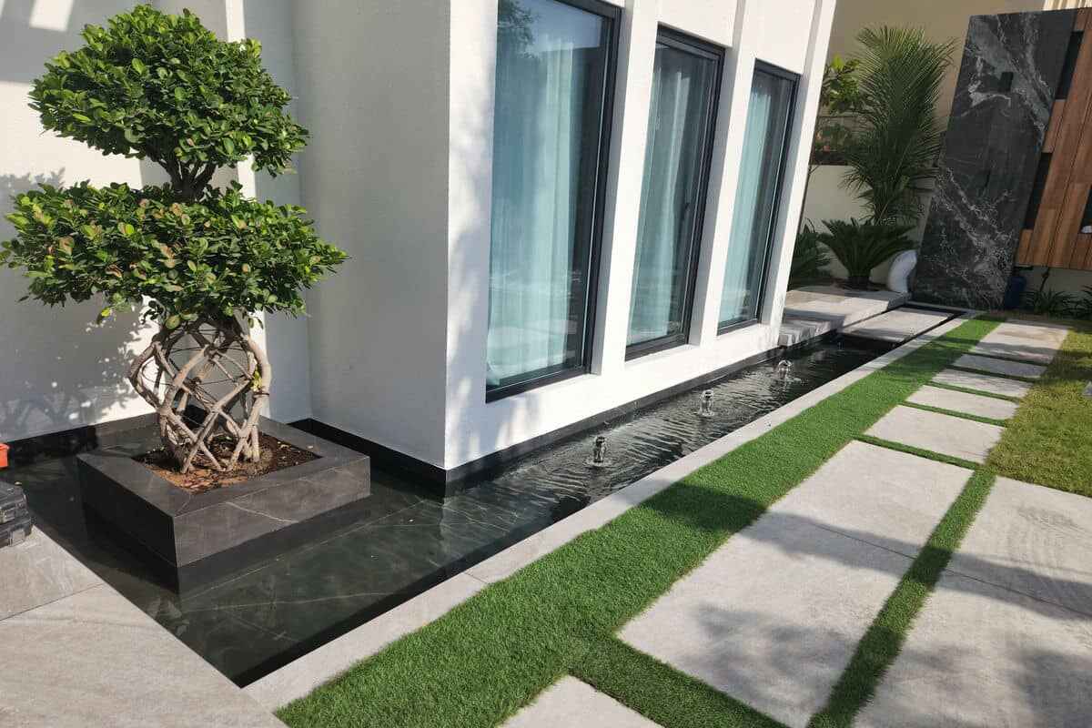 A modern exterior of a building with tall windows. In front, a rectangular planter holds a twisted-trunk tree. Adjacent to the building is a narrow water feature with small fountains, perfect for Dubai Hills living. The path is paved with white stones interspersed with green turf. Best Hard and Soft Landscaping in Dubai