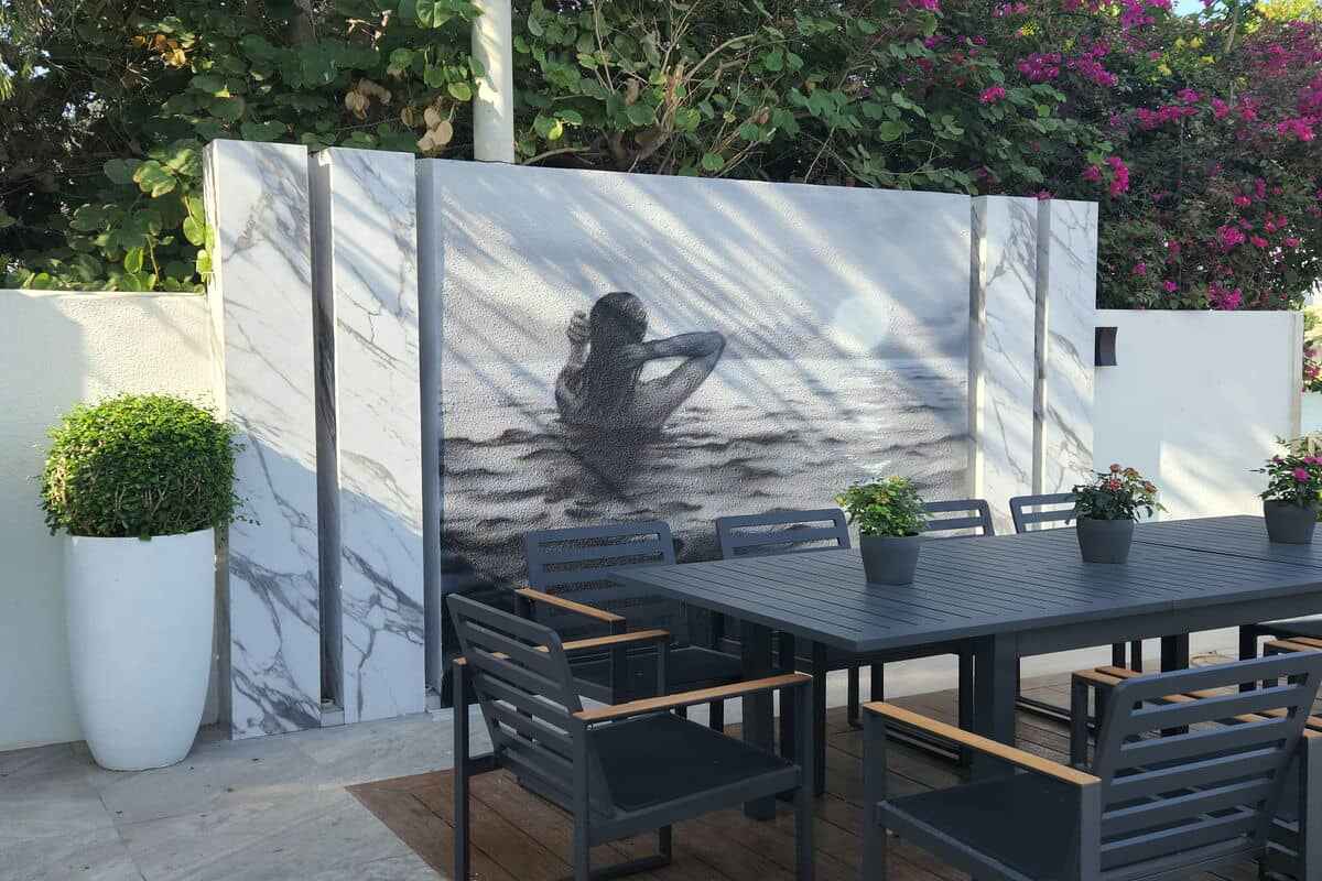 Outdoor patio featuring a dining table and chairs set on a wooden deck. Behind the table is a large, marble-like mural of a person in dark water, partially submerged. The beautifully renovated space is decorated with potted plants, and lush greenery with flowers enhances the background. Hard Landscaping Company in Dubai