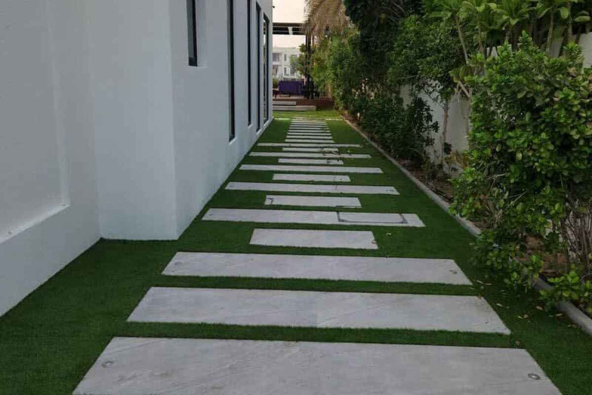 A path made of large, rectangular concrete slabs set in green lawn runs alongside a modern white building. The path, part of a recent landscaping Dubai project, is bordered by lush green bushes and trees.