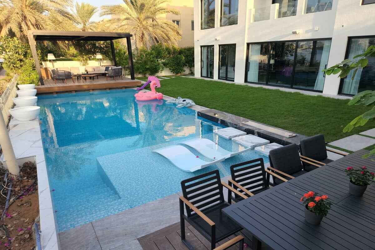 A modern backyard features a swimming pool with white submerged loungers and inflatable floaties, including a pink flamingo. The area includes a wooden deck with outdoor seating, a black dining table with flowers, and is adjacent to a contemporary house with large windows. Ideal for any Landscaping Dubai project.